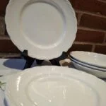 En stak hvide, flade Hanley Ware 'Satin White' tallerkener med elegant reliefkant, en står oprejst, foran murstensvæg.