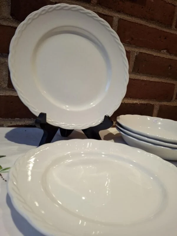 En stak hvide, flade Hanley Ware 'Satin White' tallerkener med elegant reliefkant, en står oprejst, foran murstensvæg.
