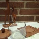 Et vintage teaktræssennepssæt med glasindsats, trælåg og en lille træske, placeret foran to teaktræsrådyrfigurer på hvid dug med broderi og murstensvæg i baggrunden.