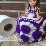 Dukke med lilla og hvidt hæklet skørt med blomsterdetaljer står ved en toiletrulle.