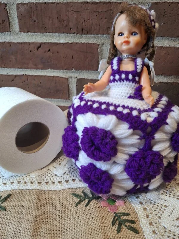 Dukke med lilla og hvidt hæklet skørt med blomsterdetaljer står ved en toiletrulle.