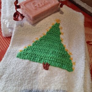 Et hvidt håndklæde med et hæklet grønt juletræ og guldstjerner, samt et stykke lyserødt roseduftpuds. Ved murstensvæg.