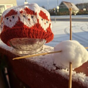 En strikket rød og hvid nissehue med julemandsansigter, placeret på et glasfad i et vinterlandskab med sne.