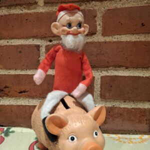 En retro julenisse figur med rød beklædning og hvidt skæg, siddende på en terracotta sparegris.