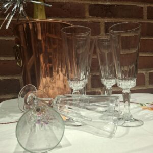Et arrangement af ni vintage champagneglas (18,5 cm høje) foran en kobberfarvet champagnekøler, en champagneflaske og en murstensvæg i baggrunden. Tre glas er væltet.