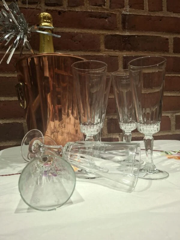Slanke champagneglas står og ligger på en hvid dug foran en kobberfarvet køler og en murstensvæg.