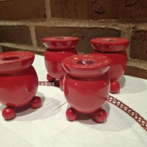 Nærbillede af fire små, røde drejede trælysestager med synlig patina til kærtelys, stående på en hvid dug foran en murstensvæg.