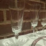 Et klart, slankt champagneglas med dekorative detaljer ved kummens bund og på stilken; flere glas står langs murstensvæggen bagved.