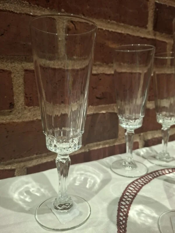 Et klart, slankt champagneglas med dekorative detaljer ved kummens bund og på stilken; flere glas står langs murstensvæggen bagved.