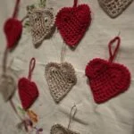En samling af håndhæklede julehjerter i rød og cremefarvet, hængende eller liggende på en hvid dug med broderier, med varierende skarphedsdybde.