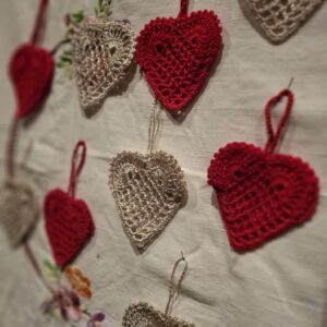En samling af håndhæklede julehjerter i rød og cremefarvet, hængende eller liggende på en hvid dug med broderier, med varierende skarphedsdybde.