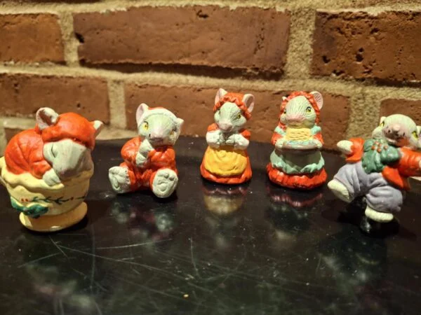 Fem musselmalede keramikfigurer forestiller en musefamilie i forskellige juleoutfits og positurer, på sort baggrund.