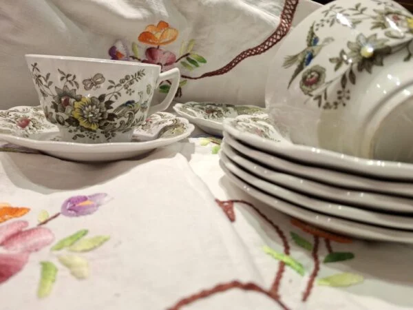 Lys Windsor-kop og underkop med farvelagt blomster- og fuglemønster på en broderet dug.