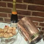 Et glas opsatsfad fyldt med småkager, der står ved siden af en champagneflaske og en kobberiskøler på en hvid dug.