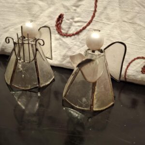 To små englefigurer lavet af glas og metal, hvor den ene holder en harpe, stående på et mørkt, reflekterende underlag.