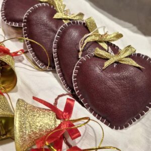Fire håndsyede bordeaux læderjulehjerter med hvide stikninger og små gyldne sløjfer, liggende på en hvid dug.