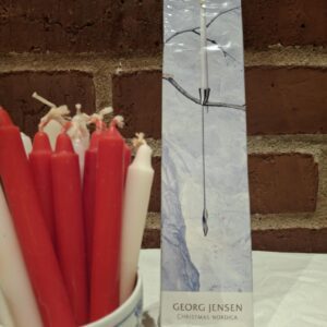 En hvid Georg Jensen Christmas Nordica lyseholder i rustfrit stål, pakket i sin originale æske, står omgivet af røde og hvide lys.