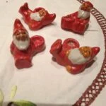 Fire små røde ler-nisser med hvide skæg, der hver især er formet til at holde et kærtelys, arrangeret på en hvid dug.