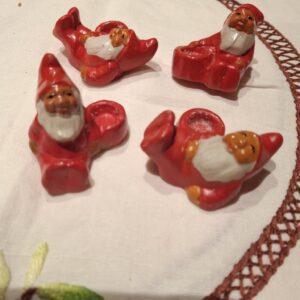 Fire små røde ler-nisser med hvide skæg, der hver især er formet til at holde et kærtelys, arrangeret på en hvid dug.