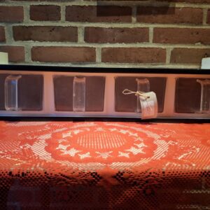 En vintage køkkenhylde med tre gennemsigtige plastskuffer, mærket 'Mel', 'Sukker' og 'Salt', monteret på en murstensvæg og stående på en rød dug.