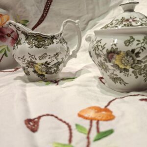 Et engelsk porcelænssæt bestående af flødekande og sukkerskål i hvid med et mønster af blomster og fasaner, stående på en hvid dug.