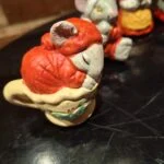 Nærbillede af en musselmalet keramikmusefigur, der sover i en gul kop med julepynt. Musen bærer en rød hue.