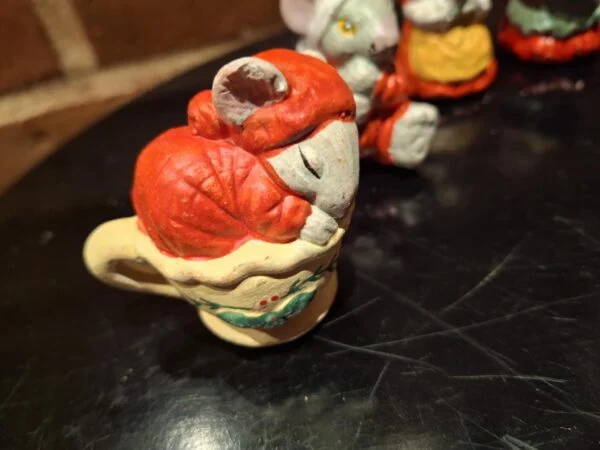 Nærbillede af en musselmalet keramikmusefigur, der sover i en gul kop med julepynt. Musen bærer en rød hue.