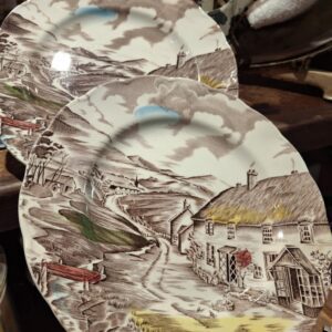 To engelske fajancetallerkener 'Quiet Day' med et malerisk landskabsmotiv af en landsby og hytte, domineret af brune og grå farver.