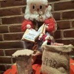 Siddende nisse med briller, hvidt skæg, lille pibe og bog i flettet stol. Foran står en skammel og en julesæk med små dekorationer.