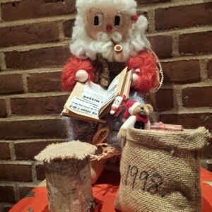 En stor håndlavet nisse med et hvidt skæg, briller og nissehue, læsende en bog. Ved siden af en julesæk og gaver.