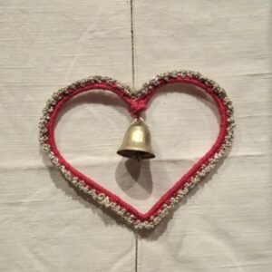 Tre røde, hæklede julehjerter, hver med en lille messingklokke hængende i midten, arrangeret lodret på en hvid dug.