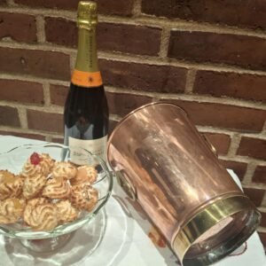 En skinnende vintage champagnekøler i kobber og messing (22 cm høj), placeret skråt ved siden af en champagneflaske og en glasskål med småkager på en hvid dug foran en murstensvæg.