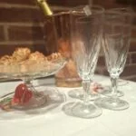 To slanke champagnefløjter med snoet mønster på en hvid dug, med kagefad, flaske og kobberfarvet køler i baggrunden.