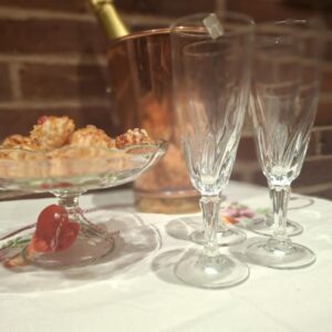 Fire elegante champagnefløjter med fleur-de-lis-mønster i bunden, stående ved siden af en kobberfarvet champagnekøler og en skål med små kager, alt på en hvid dug foran en murstensvæg.