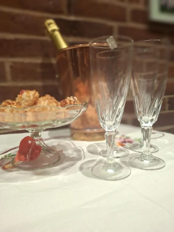 To slanke champagnefløjter med snoet mønster på en hvid dug, med kagefad, flaske og kobberfarvet køler i baggrunden.