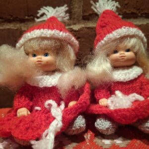 To nissepiger med strikkede røde kjoler, hvide kraver og huer med kvast. De har blondt hår og blå øjne.