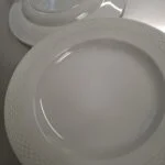 To hvide Aluminia tallerkener med en flettet kant, den ene ovenpå den anden, på en reflekterende overflade.