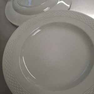 To hvide Aluminia tallerkener med en flettet kant, den ene ovenpå den anden, på en reflekterende overflade.