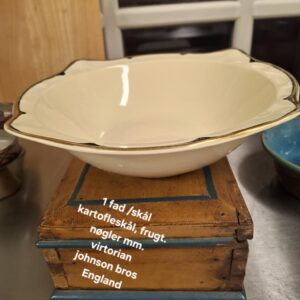 Oval cremefarvet Johnson Bros. 'Virton' skål med elegant guld- og sort kant, placeret på en rustik træboks. Teksten '1 fad /skål kartoffelskål, frugt. nøgler mm. virtorian johnson bros England' er synlig.