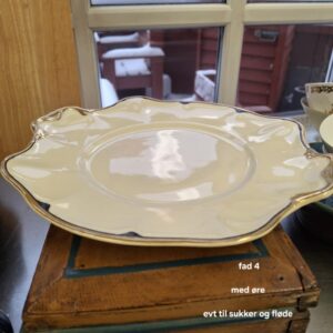 Et antikt, cremefarvet fad med guldkant, bølget kant og to 'ører', placeret ovenpå en rustik træboks med udsigt til et snedækket landskab i baggrunden. Teksten 'fad 4 med øre 26 x 24 cm virtorian johnson bros England' står på billedet.