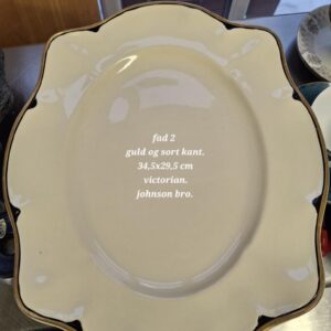 Elegant, ovalt, cremehvidt Johnson Bros. fad (Victorian) med bølget kant og en detaljeret guld- og sort bort. Teksten 'fad 2 guld og sort kant. 34,5x29,5 cm victorian. johnson bro.' er synlig.