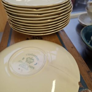 Stak af 11 cremehvide underkopper med guldkant fra Johnson Bros. 'Victorian'. Underkopperne er stablet på en rustik træoverflade med blå detaljer. Bagsiden af den øverste underkop har et stempel.