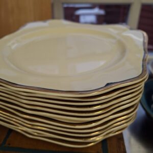 Stabel af 12 cremefarvede, antikke frokosttallerkener fra Johnson Bros. 'Victoria' med guldbort og blå streg, set skråt fra siden. Teksten '12 Frokost med guld Victoria Johnson bro England' er synlig.