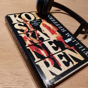 Bog Korsikaneren af William Heffernan med sort omslag og cremefarvet titeltekst, hovedtelefoner liggende på den