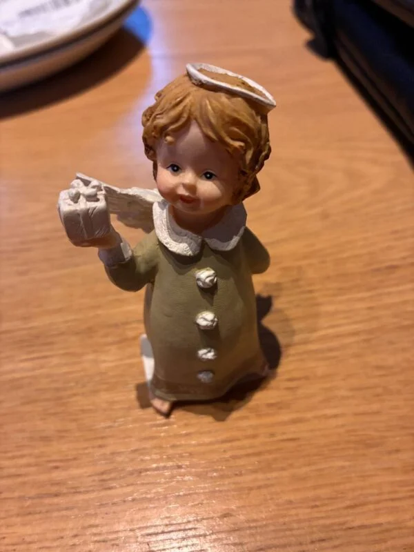 Englefigur i grønlig kjole med en gave i hånden og et synligt skår i den lyse glorie.