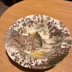 Morgenmadsskål med et gammelt hus, små figurer og brun bladranke langs kanten.