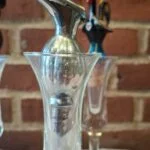 Sølvfarvet skænketud med låg og synlig patina, placeret i et separat tulipanformet glas.