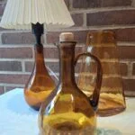 Ravfarvet bordlampe, karaffel med korkprop og en høj vase fotograferet sammen