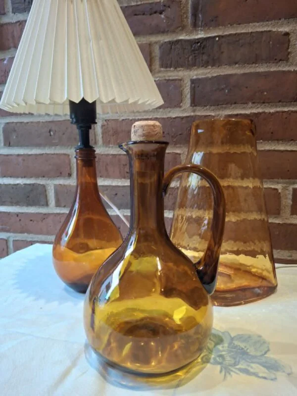 Ravfarvet bordlampe, karaffel med korkprop og en høj vase fotograferet sammen
