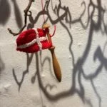 Håndhæklet rød-hvidt Dannebrog på en kniplepind i træ, hængt i rød snor på en snoet gren.